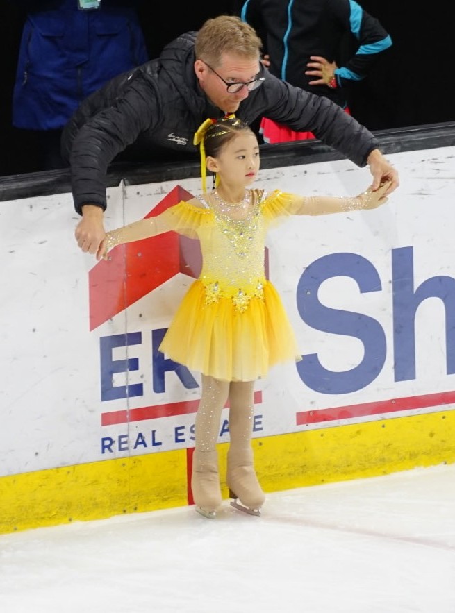 Coach Nick guiding a young skater in a yellow dress at the rink boards
