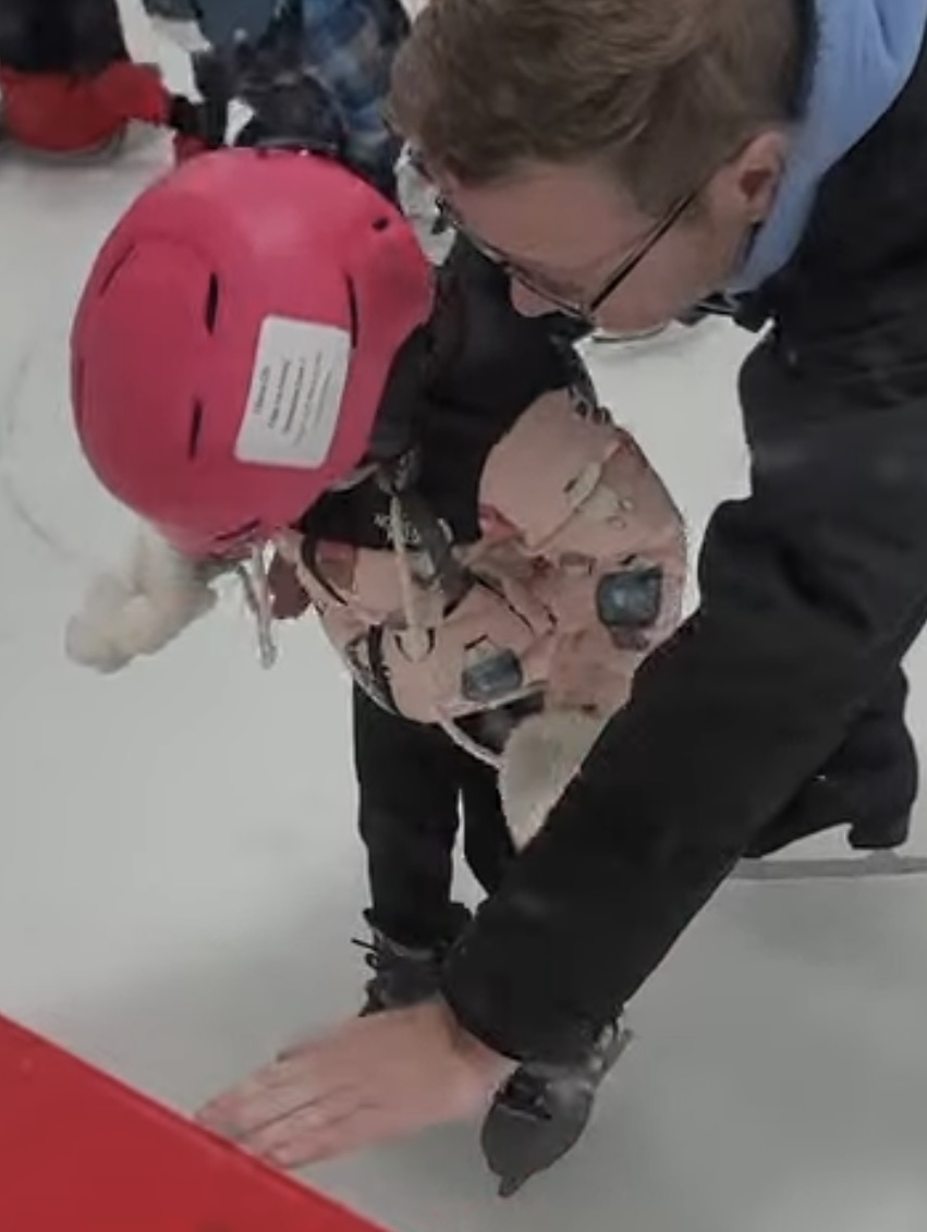 Coach Nick helping a young beginner wearing a pink helmet on the ice